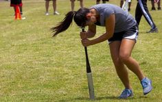 a young woman holding a baseball bat on top of a lush green field with other people in the background