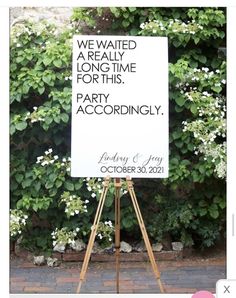 a white sign sitting on top of a wooden easel in front of a lush green hedge