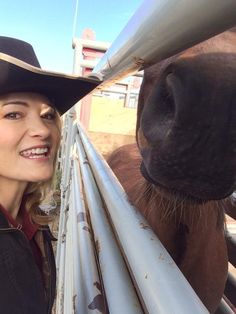 a woman wearing a cowboy hat standing next to a brown horse behind a metal fence