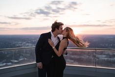 a man and woman kissing on top of a building with the sun setting behind them