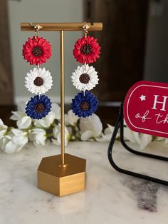 two red, white and blue flower dangle earrings on a stand with flowers in the background