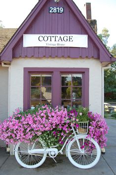 a white bicycle with flowers in the basket parked next to a purple building that says cottage