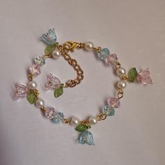 This bracelet is 7.5 inches long with a 2-inch extender. It has pink and blue flower charms with green leaf accents, clear beads, and faux pearls, all connected by a gold chain. It fastens with a lobster clasp. Floral Beaded Bracelet, Fairycore Bracelet, Fairy Bracelet, Fairy Bracelets, Clear Bracelet, Green Beaded Bracelets, Diy Collier, Clear Beads, Leaf Bracelet