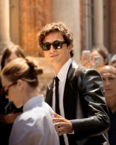 a man wearing sunglasses and a black leather jacket walking past a woman in a white dress