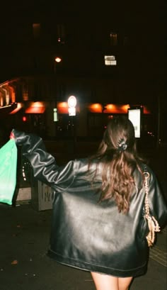 a woman in a black leather jacket is walking down the street with a green bag