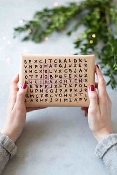 two hands holding up a box with letters on it and christmas greenery in the background