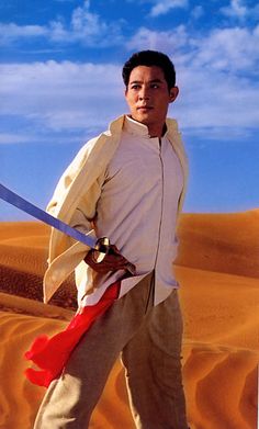 a man holding two swords standing in the desert with sand dunes behind him and blue sky