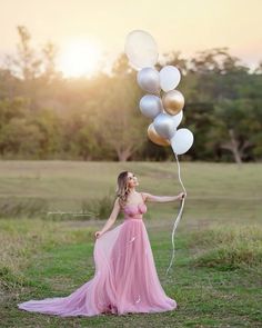 a woman in a pink dress holding some white and gold balloons with the sun setting behind her