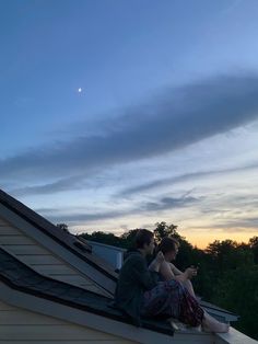 two people sitting on top of a roof looking at their cell phones while the sun is setting