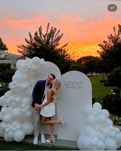 a man and woman kissing in front of a heart - shaped arch with balloons at sunset