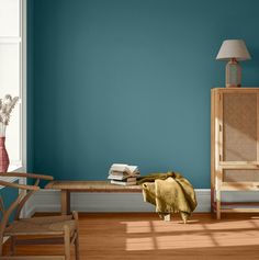 a living room with blue walls and wooden furniture in the foreground is a table, chair, lamp, and bookshelf