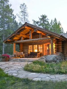 a log cabin is lit up at night in the woods with patio furniture and stone walkway