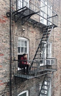 a person sitting on a window sill next to a fire escape ladder in front of a brick building