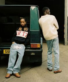 two people sitting on the back of a green car in front of a black van