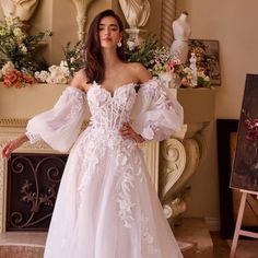 a woman standing in front of a fireplace wearing a white wedding dress with long sleeves