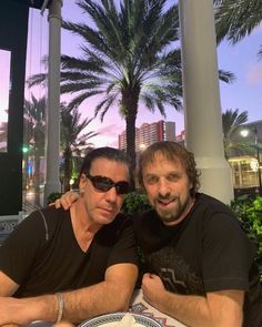 two men sitting at an outdoor table with plates and drinks in front of palm trees