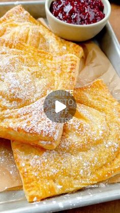 crepes with powdered sugar on top sit in a tray next to a bowl of jam