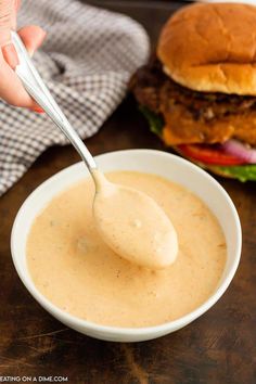 a person holding a spoon over a bowl of soup with a hamburger in the background