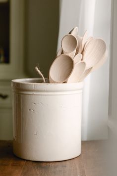 several wooden spoons in a white container on a wood table next to a window