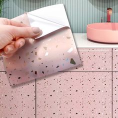 a hand holding a piece of paper over a pink counter top with a sink and mirror in the background