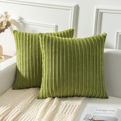 two green pillows sitting on top of a white couch next to a book and vase