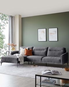 Modern charcoal grey sectional sofa with clean lines and deep, comfortable cushions, complemented by a sleek black metal frame and legs. A matching, elegant grey throw blanket is draped over one side, with an assortment of earth-toned decorative pillows adding a touch of warmth to the setup. A minimalist black rectangular coffee table with a glass top and a collection of books is positioned in front of the sofa on a textured off-white area rug. Living Room Light Grey Floor, Light Sofa Living Room Color Schemes, Contemporary Living Room Grey Couch, Living Room With Feature Wall, Living Room Light Grey Sofa, Light Green Sofa Living Room Ideas, Sofa Colors For Living Room, Light Green Living Room Walls, Light Green Living Room Color Scheme