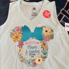 a white tank top with flowers and garden festival on it's chest is sitting on a bed