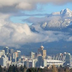 the city is surrounded by tall buildings and snow capped mountains in the distance, with clouds hovering over them