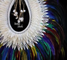 A handmade Papua New Guinea tribal inspired large white braided Rope necklace, decorated with one hundred and forty seven white Shell slices, a band of round silver beads, and a fringe of purple, yellow, green, blue, red and iridescent black dyed Feathers. The inner width of the band of the necklace is 5.25 inches, and the maximum outer width of the necklace is 23 inches. The drop length from the top of the necklace to the bottom of the feathers,is 27 inches. The white Shells are, between 1.3 an White Macrame Necklace For Festivals, White Macrame Necklace For Festival, White Bohemian Woven Jewelry, Bohemian White Woven Jewelry, White Handwoven Artisan Necklace, White Handwoven Festival Necklace, White Handwoven Necklace For Festivals, White Artisan Beaded Necklaces For Festivals, Artisan White Beaded Necklaces For Festivals