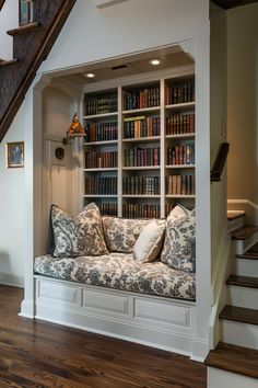 a room with a couch, bookshelf and stairs leading up to the second floor