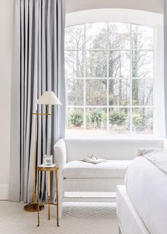 a white bed sitting under a large window next to a lamp and a table in front of it