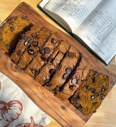 a wooden cutting board topped with slices of chocolate chip pumpkin bread next to an open book