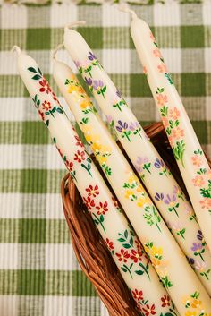 four white candles in a basket with flowers on them sitting on a checkered tablecloth