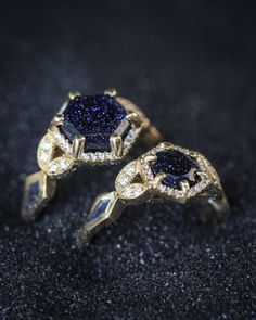 Two blue goldstone engagement rings from Staghead Designs are shown in black sand. One is our regular "Lucy In The Sky" design with a hexagon cut blue goldstone & the other is "Lucy In The Sky Petite" with a round cut blue goldstone. Both rings feature a hexagon-shaped diamond halo, floral diamond accents, & crushed blue goldstone inlays. Whiskey Barrel Wedding Ring, Antler Wedding Rings, Handcrafted Engagement Ring, Wood Wedding Ring, Alternative Wedding Rings, Blue Goldstone, Custom Ring Designs, Wedding Rings Rose Gold, Men's Wedding Ring