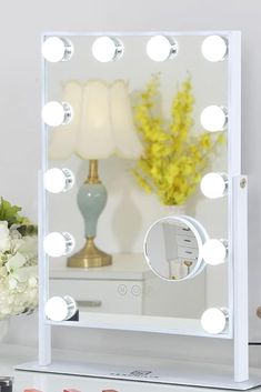 a white vanity mirror with lights on it and flowers in the corner next to it