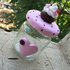 a pink cupcake in a jar with a heart on the lid sitting on a rock