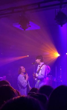 two people standing in front of a microphone on stage with purple lights shining down the room