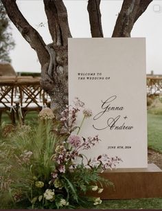 a sign that is sitting in the grass next to some flowers and trees with chairs behind it