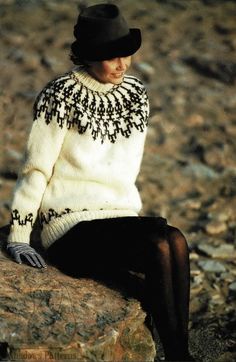 a woman sitting on top of a rock wearing a black hat and white knit sweater