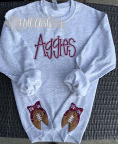 Aggie team puff embroidered sweatshirt with football side bows on bottom band of shirt. We use a Gildan brand unisex sweatshirt. These are so cute on and great for game day!!  These can be made in your team colors. Embroidered team name can be done in your choice of thread color.  The side football bows are so flattering on any body type. They open the band around the sweatshirt with the cut at the center bottom of the football. They make the waistband looser and a flattering look. Pair it with a pair of shorts, jeans, leggings and you are going to look great cheering on your Aggies!! For reference, Angie is wearing a size Medium sweatshirt. These are unisex sizing. I would suggest ordering in your normal size unless you want a bigger, comfy, oversized fit. These wash nicely. I suggest was Football Bows, Puff Embroidery, Team Sweatshirts, Jeans Leggings, Team Name, Embroidered Sweatshirt, Shorts Jeans, Team Names, Embroidered Sweatshirts