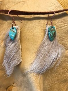 two turquoise and white stone earrings hanging from a leather cord on a tan surface with fringes