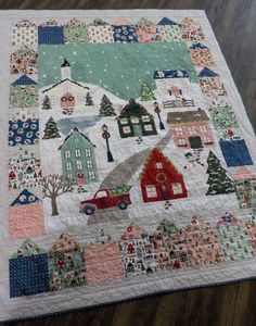 a quilted table topper with houses and trees on it, sitting on a wooden floor