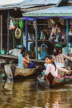 two children are sitting in small boats on the water near a shack with several other people