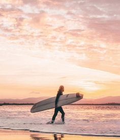 a person walking on the beach with a surfboard in their hand as the sun sets