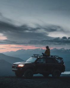 a man standing on top of a vehicle in the middle of mountains at sunset or dawn