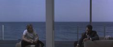 a man sitting on a couch talking on a cell phone next to the ocean while looking out at the water