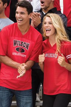 two people in red shirts standing next to each other at a basketball game with fans behind them