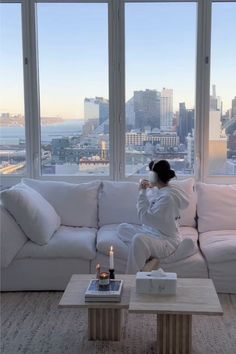 a woman sitting on top of a white couch in front of a window next to a coffee table