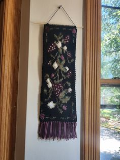 a black wall hanging with flowers and tassels on the bottom, in front of a window