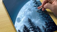 a hand is holding a paintbrush over a painting of a full moon and trees